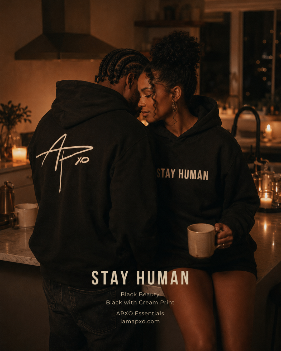 Candlelit kitchen at night: a couple in matching Stay Human Heavyweight Hoodies in Black Beauty, one showing the cream APXO back signature.