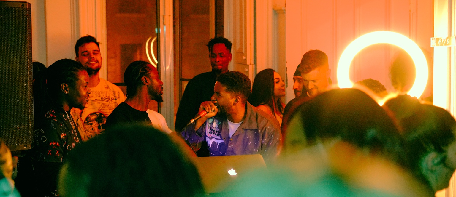 APXO performing with a microphone in a crowded room bathed in warm orange light