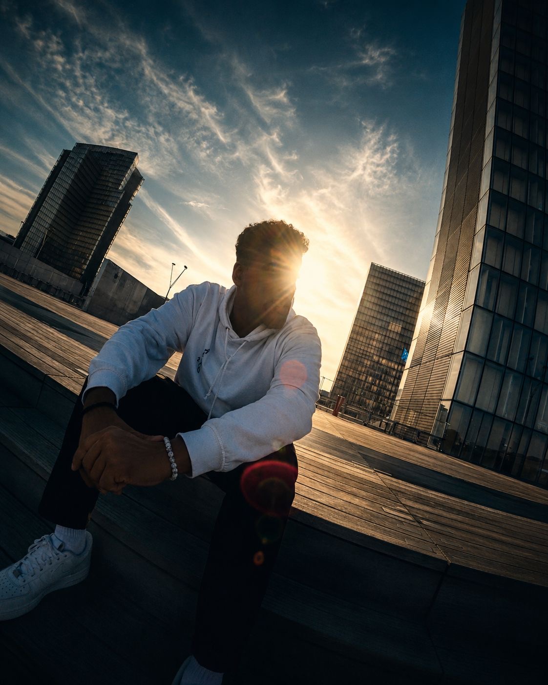 APXO seated on wide outdoor steps at sunset with tall buildings behind him