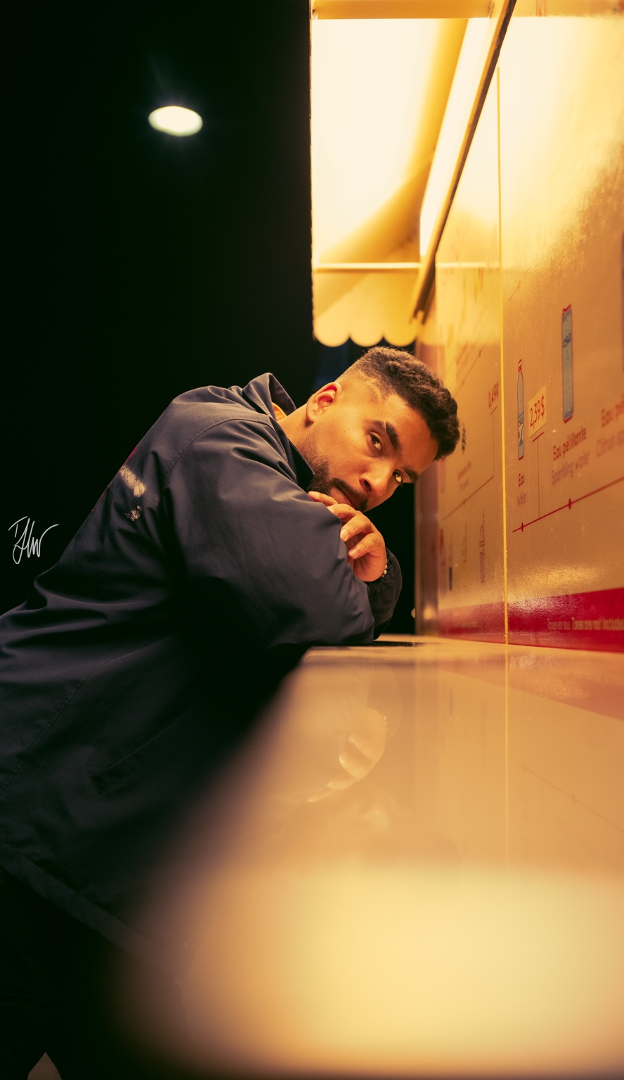 APXO leaning against a warm-lit counter at night looking towards the camera