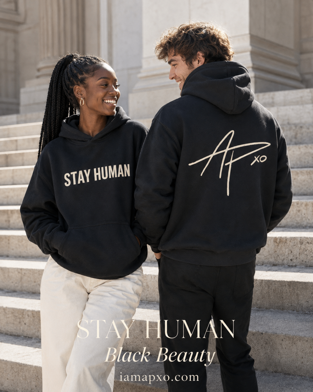 Fashion editorial on stone steps: a woman in the Stay Human Heavyweight Hoodie facing camera and a man turned away showing the cream APXO signature on the back, black with cream print.