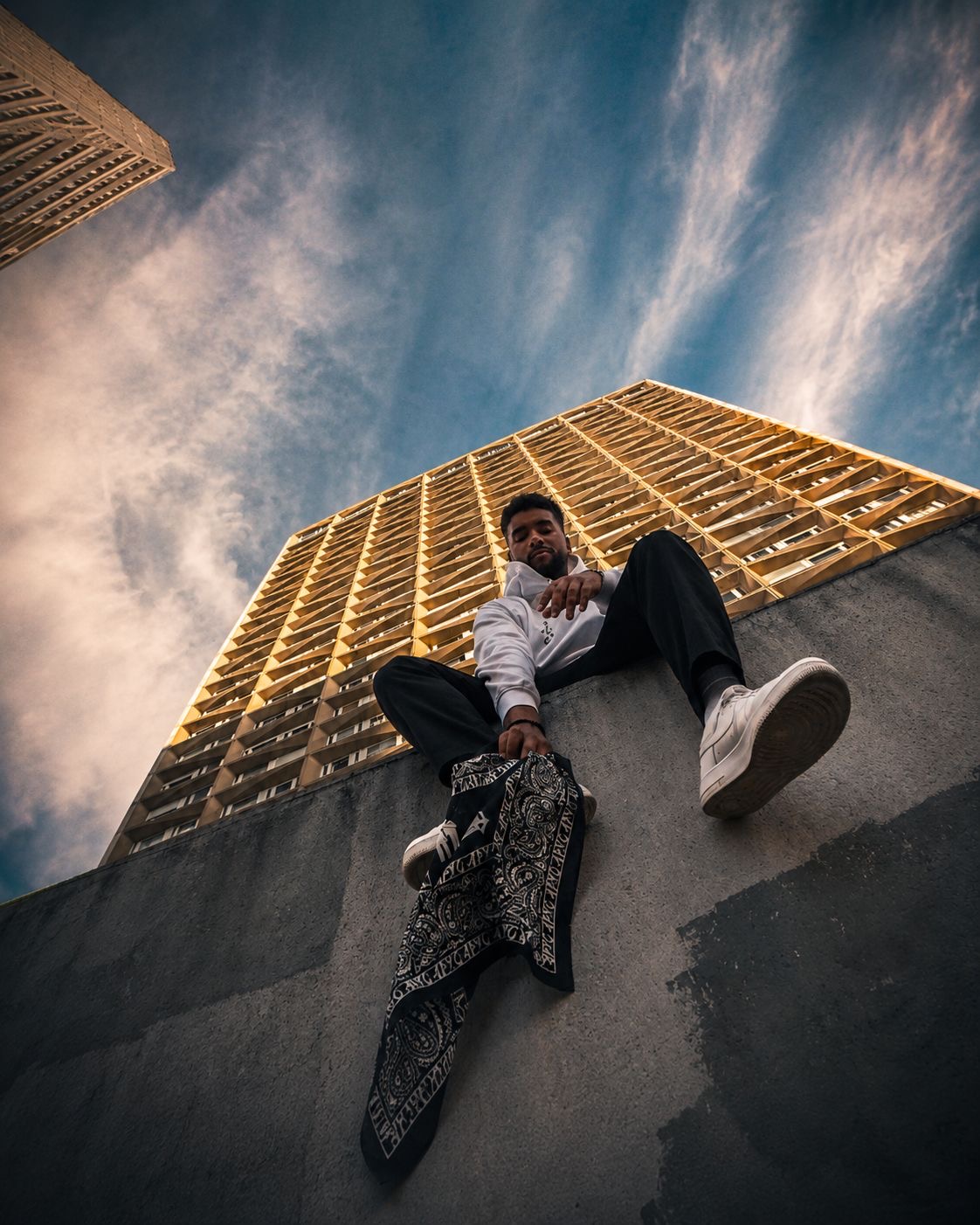 APXO seated on a ledge in front of a modern building holding a bandana