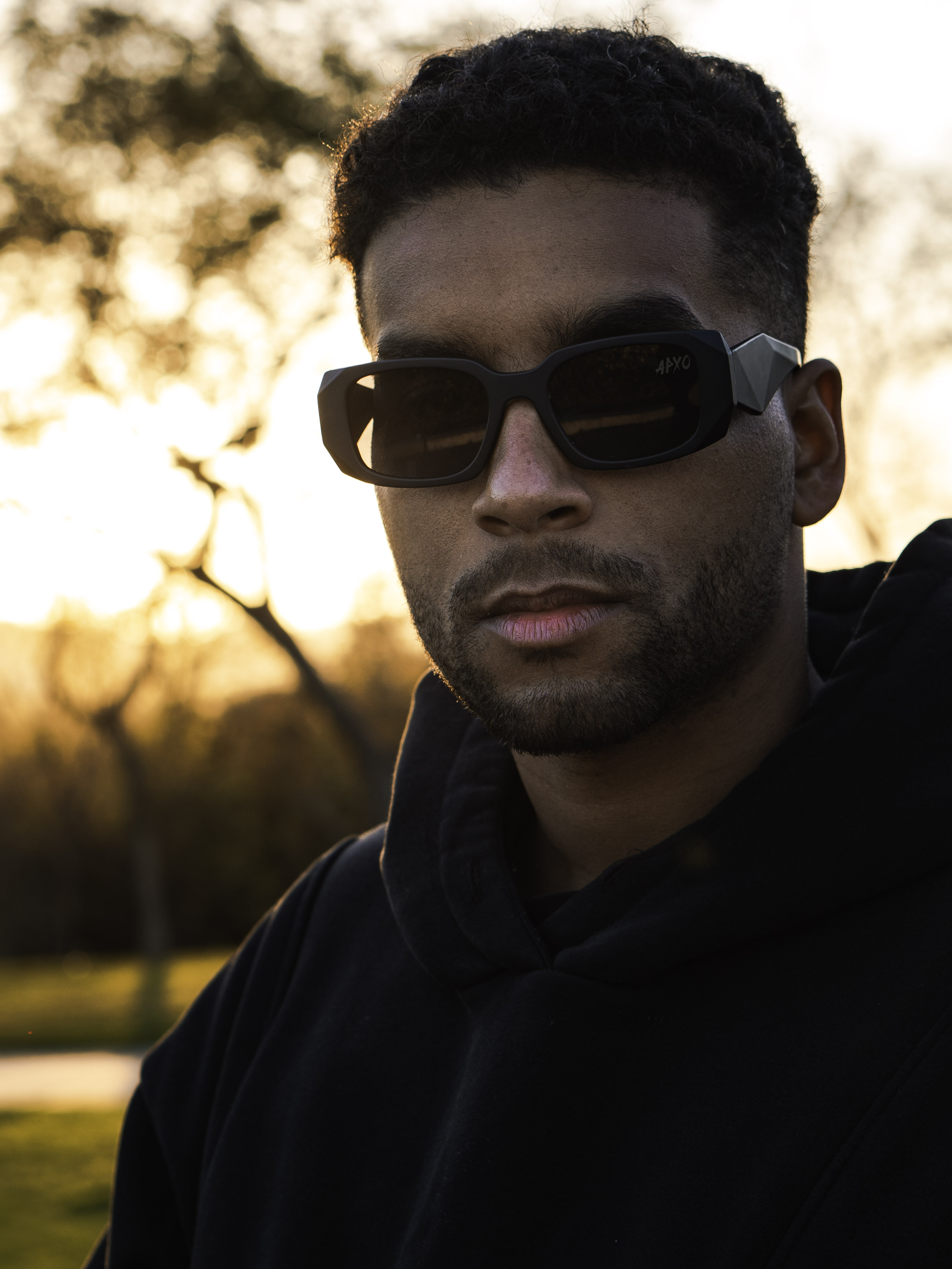 Golden hour portrait of APXO wearing dark APXO sunglasses and a black hoodie