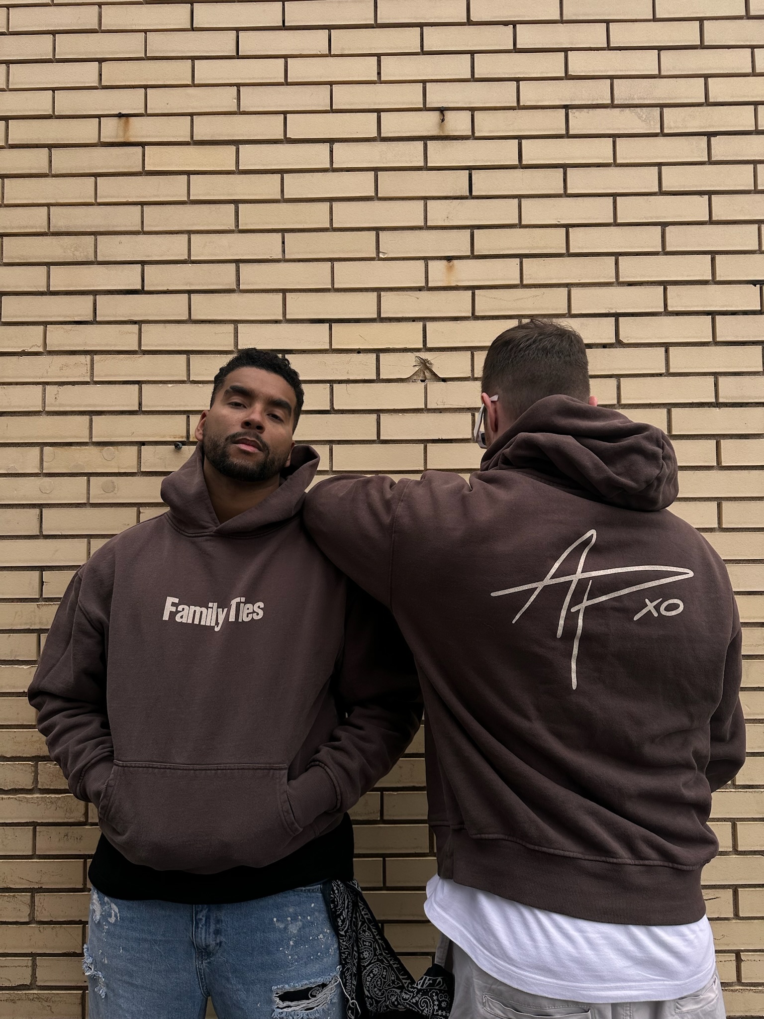Two figures against a brick wall wearing brown Family Ties and APXO hoodies